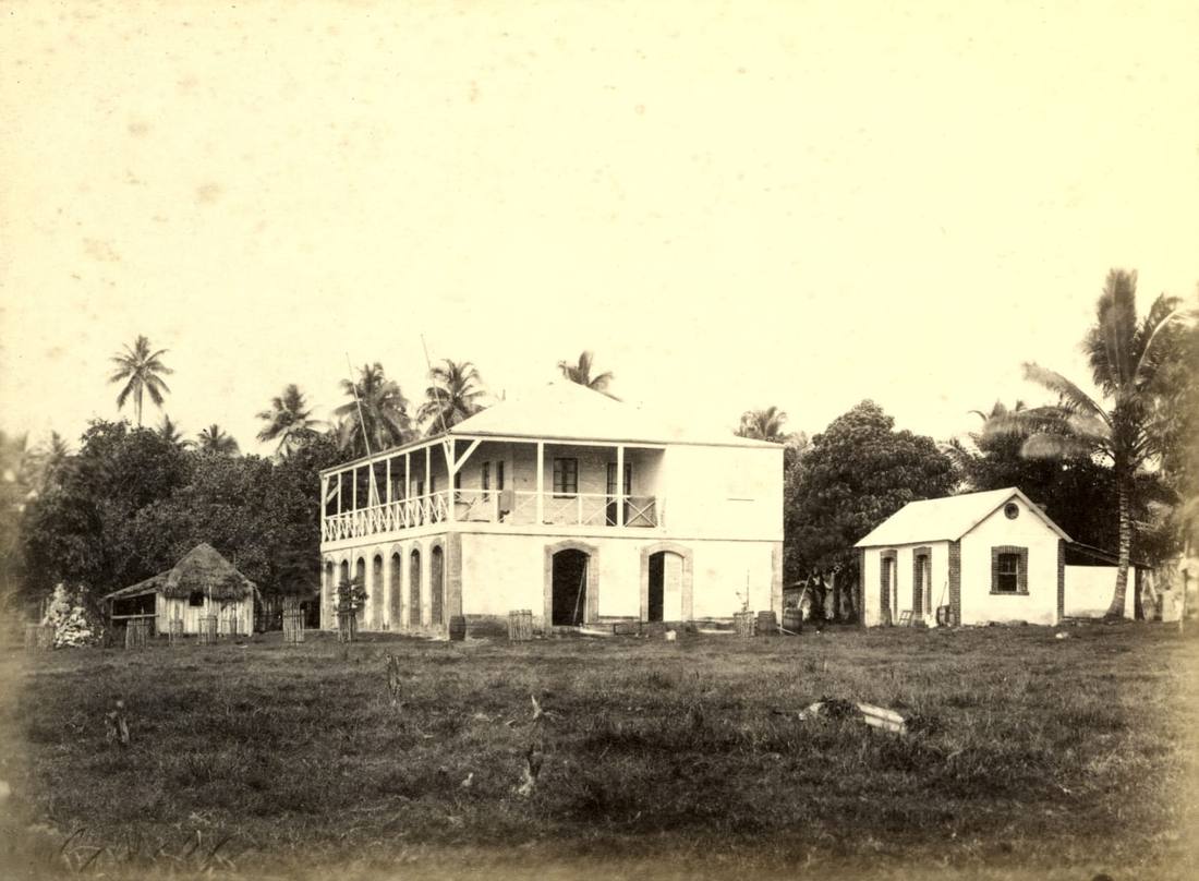 A two-storey trading house with a wide upstairs verandah and arched doorways, a small brick building and a thatched hut beside it, coconut palms behind, 1886